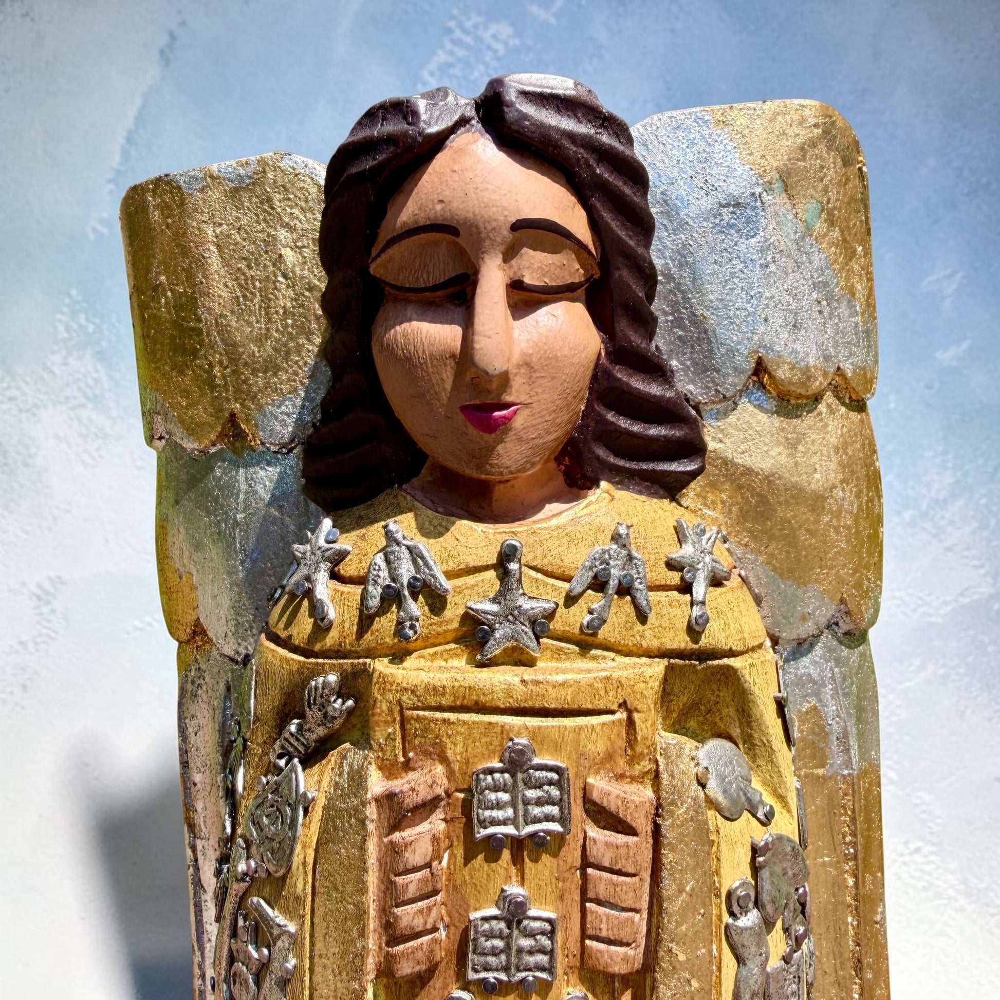 Milagros wooden guardian angel figurine wearing a yellow robe with gold and silver metallic details, standing on a light blue textured background, close-up of Angel’s face.