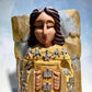 Milagros wooden guardian angel figurine wearing a yellow robe with gold and silver metallic details, standing on a light blue textured background, close-up of Angel’s face.
