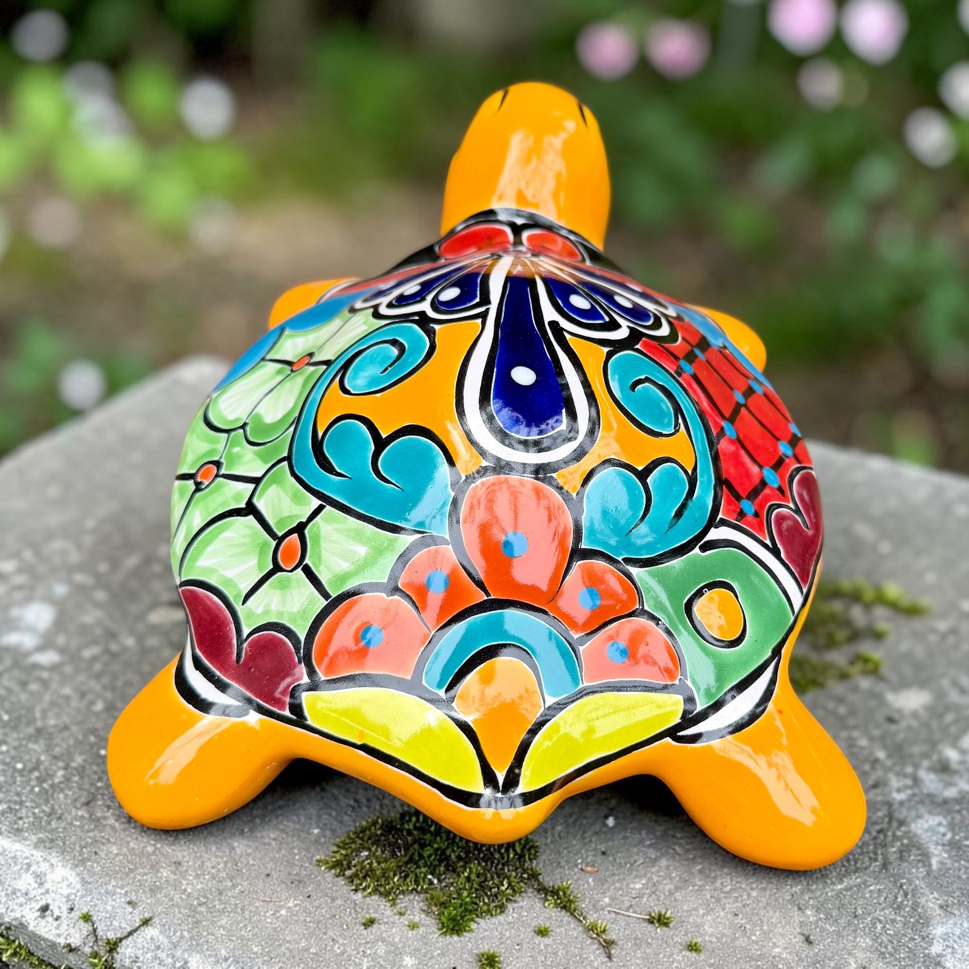 A glossy ceramic turtle figurine with a bright yellow body and a highly decorated shell in red, blue, yellow, black, and white, resting on a stone surface with a blurred garden background, back shell view.