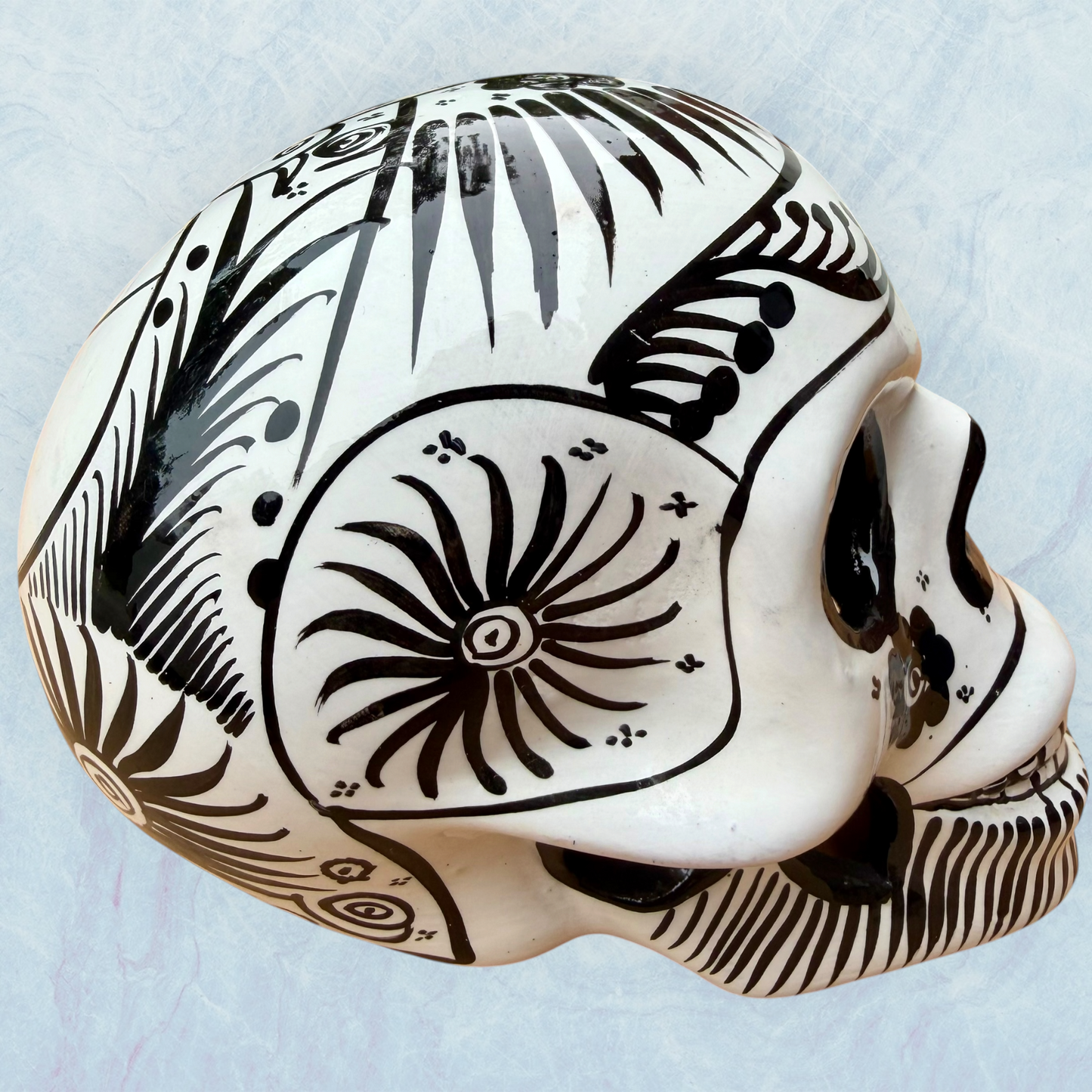 A decorative skull sculpture with intricate black sugar skull style patterns painted on an off-white base, set against a light blue textured background, left side view.