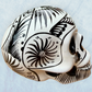A decorative skull sculpture with intricate black sugar skull style patterns painted on an off-white base, set against a light blue textured background, left side view.