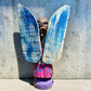 A handmade wood Milagros angel figurine with large gold wings, holding a guitar, dressed in a colorful patterned robe against a textured blue wall. Back view with hanging loop.