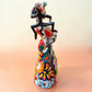 A Talavera Day of the Dead (Día de los Muertos) Frida figurine  with a monkey on her shoulder and a red parrot on her other shoulder, set against a light blue gradient background. Left side view of figurine.