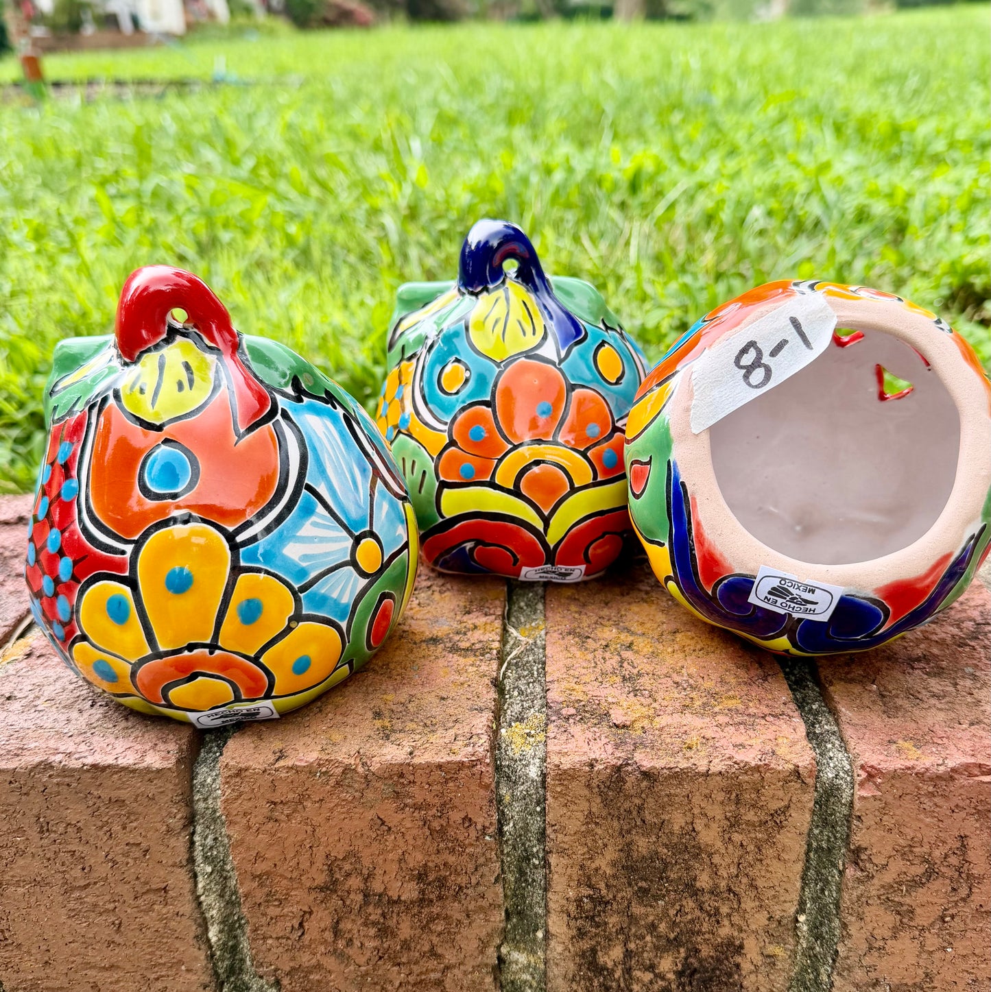 Three hand-painted Talavera ceramic jack-o'-lantern pumpkins in bright orange, yellow, red, green, and blue, sitting on a brick ledge with a blurred green lawn and house in the background, bottom opening visible.