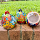 Three hand-painted Talavera ceramic jack-o'-lantern pumpkins in bright orange, yellow, red, green, and blue, sitting on a brick ledge with a blurred green lawn and house in the background, bottom opening visible.