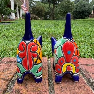 Two colorful hand-painted talavera ceramic cat figurines in cobalt blue with multi color designs standing on a brick path with a green lawn back view with pointy tail.