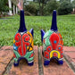 Two colorful hand-painted talavera ceramic cat figurines in cobalt blue with multi color designs standing on a brick path with a green lawn back view with pointy tail.