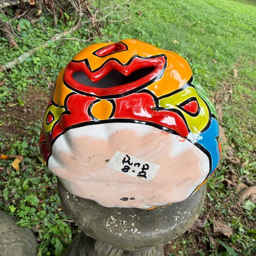A hand-painted ceramic jack-o'-lantern pumpkin ornament with a bright orange face, red carved eyes and mouth, a blue stem, and colorful side patterns, sitting outdoors on a stone pedestal in a grassy yard.  This show the bottom with the item number.