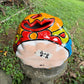A hand-painted ceramic jack-o'-lantern pumpkin ornament with a bright orange face, red carved eyes and mouth, a blue stem, and colorful side patterns, sitting outdoors on a stone pedestal in a grassy yard.  This show the bottom with the item number.