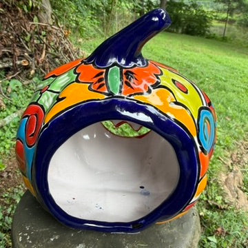 A hand-painted ceramic jack-o'-lantern pumpkin ornament with a bright orange face, red carved eyes and mouth, a blue stem, and colorful side patterns, sitting outdoors on a stone pedestal in a grassy yard. This is the back view with the opening for a battery candle.