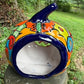 A hand-painted ceramic jack-o'-lantern pumpkin ornament with a bright orange face, red carved eyes and mouth, a blue stem, and colorful side patterns, sitting outdoors on a stone pedestal in a grassy yard. This is the back view with the opening for a battery candle.