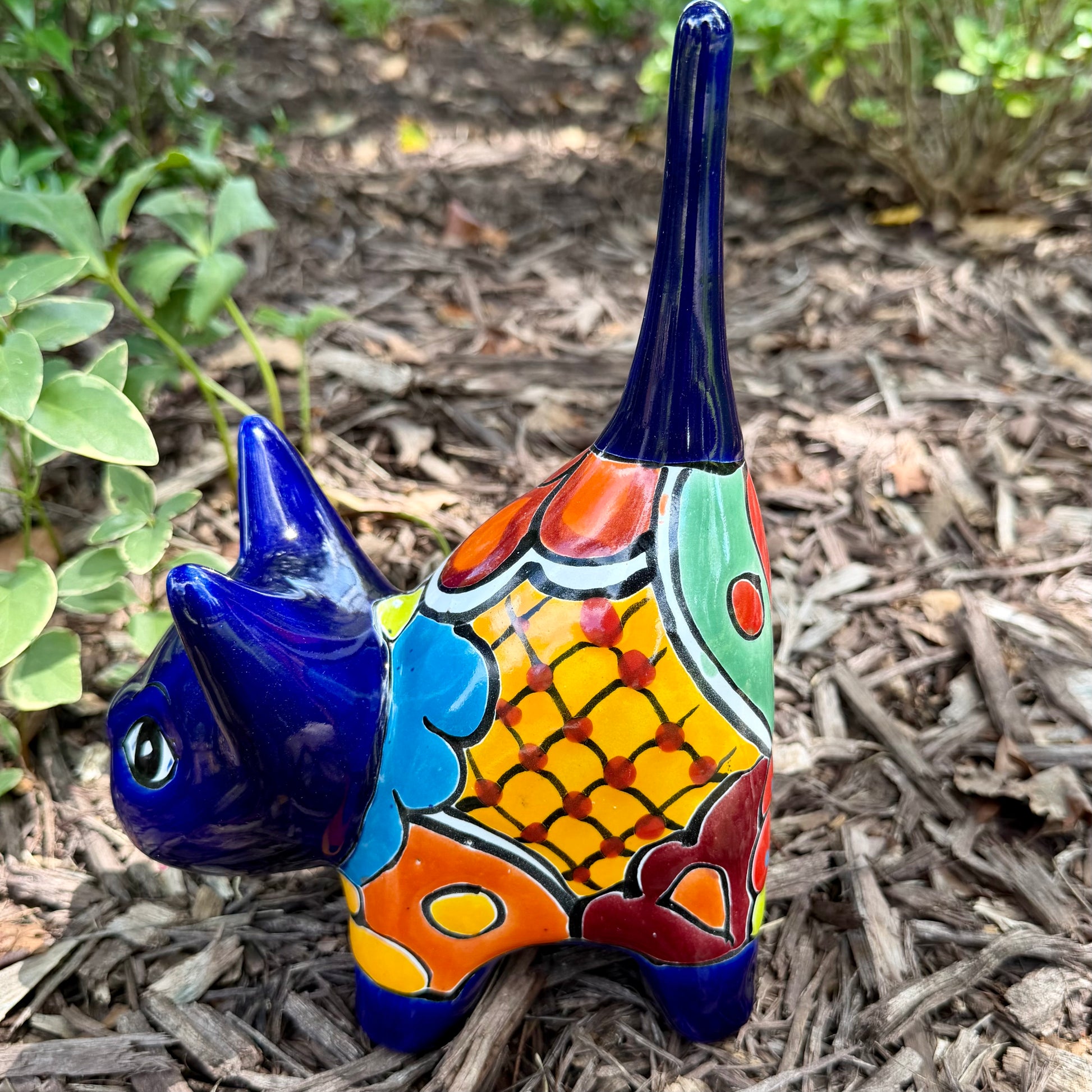 A colorful glazed ceramic cat figurine standing on mulch outdoors, with green plants blurred in the background. The cat is decorated with blue, orange, yellow, and white patterns, right side view.