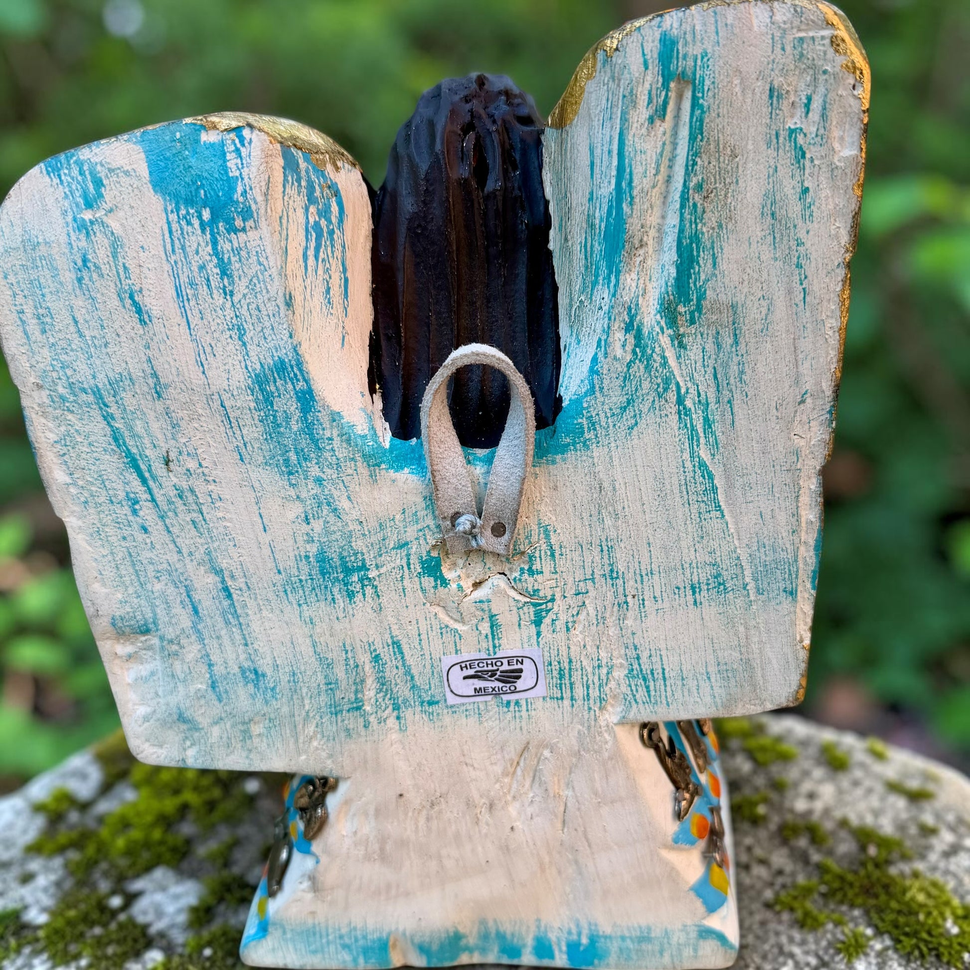 A Milagros guardian angel figurine with gold wings, dark hair, and a blue, white, and gold embroidered dress, posed outdoors on a stone surface with a blurred green background. This is the back view showing handing loop