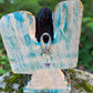 A Milagros guardian angel figurine with gold wings, dark hair, and a blue, white, and gold embroidered dress, posed outdoors on a stone surface with a blurred green background. This is the back view showing handing loop