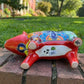 A colorful hand-painted red Talavera ceramic pig figurine standing on a brick surface with a blurred green lawn and trees in the background, bottom view with drain hole.