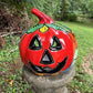 A hand-painted talavera Halloween  jack-o'-lantern candle holder in red, green, black, and orange, displayed on a stone pedestal in an outdoor garden setting.