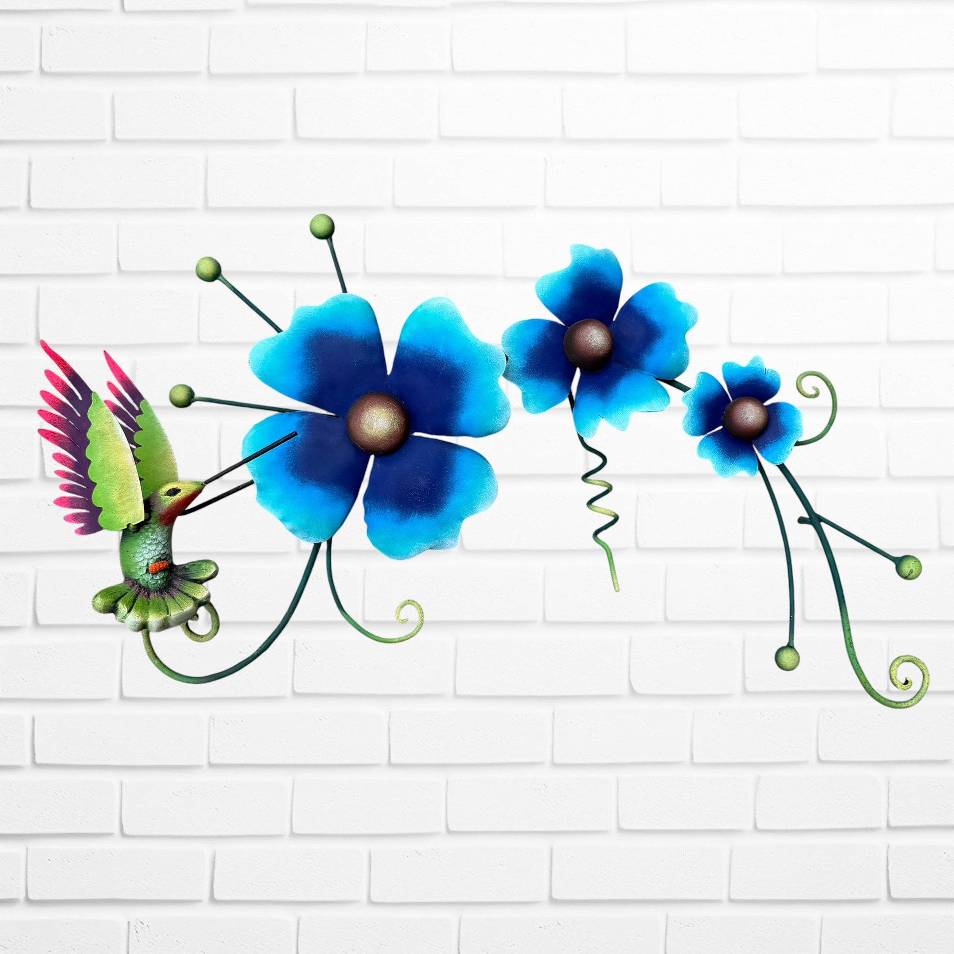 Decorative metal wall art with three blue flowers and a colorful hummingbird on a white brick background