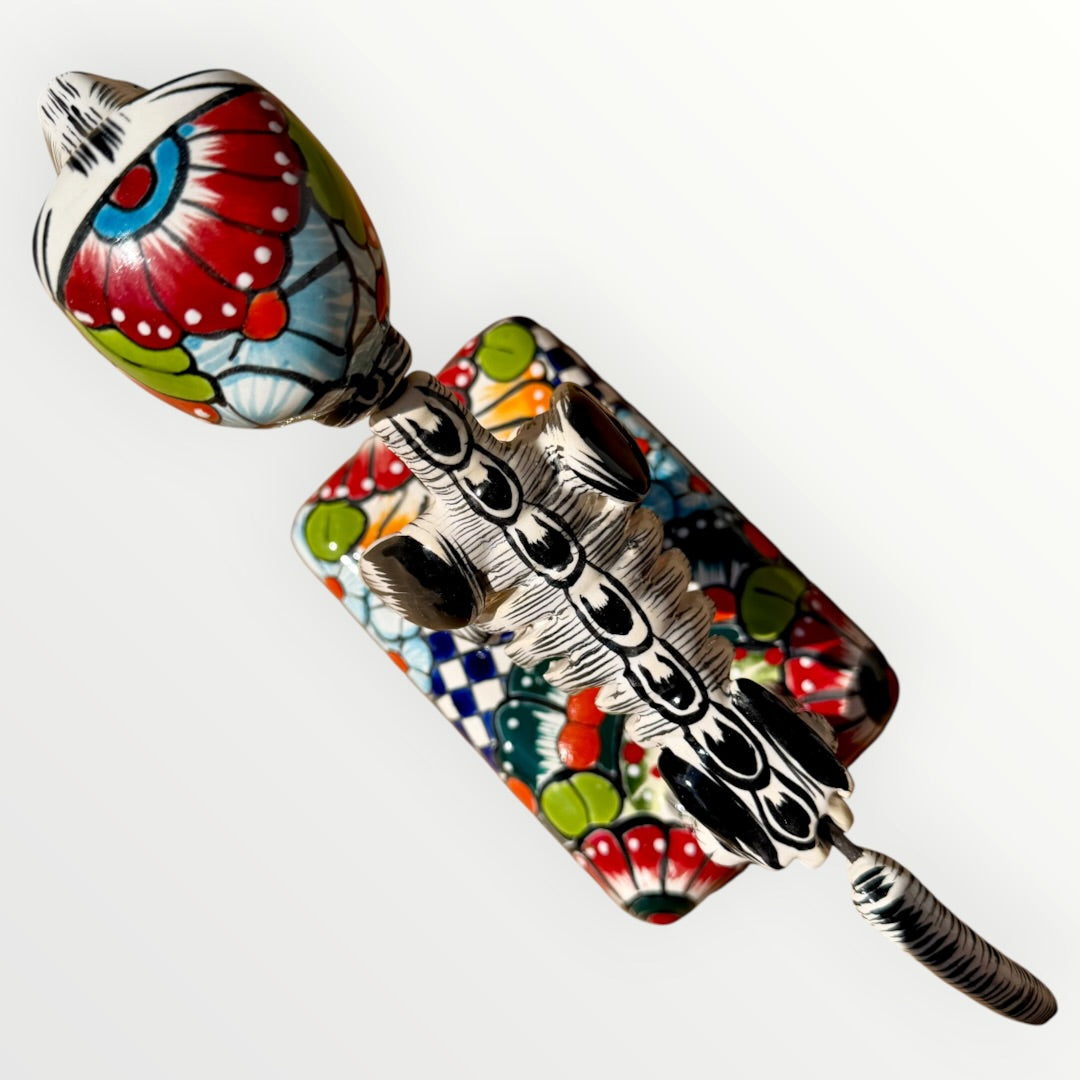 A ceramic Mexican-style skeleton dog figurine standing on a colorful floral base against a plain white background. Top view of catrina dog.