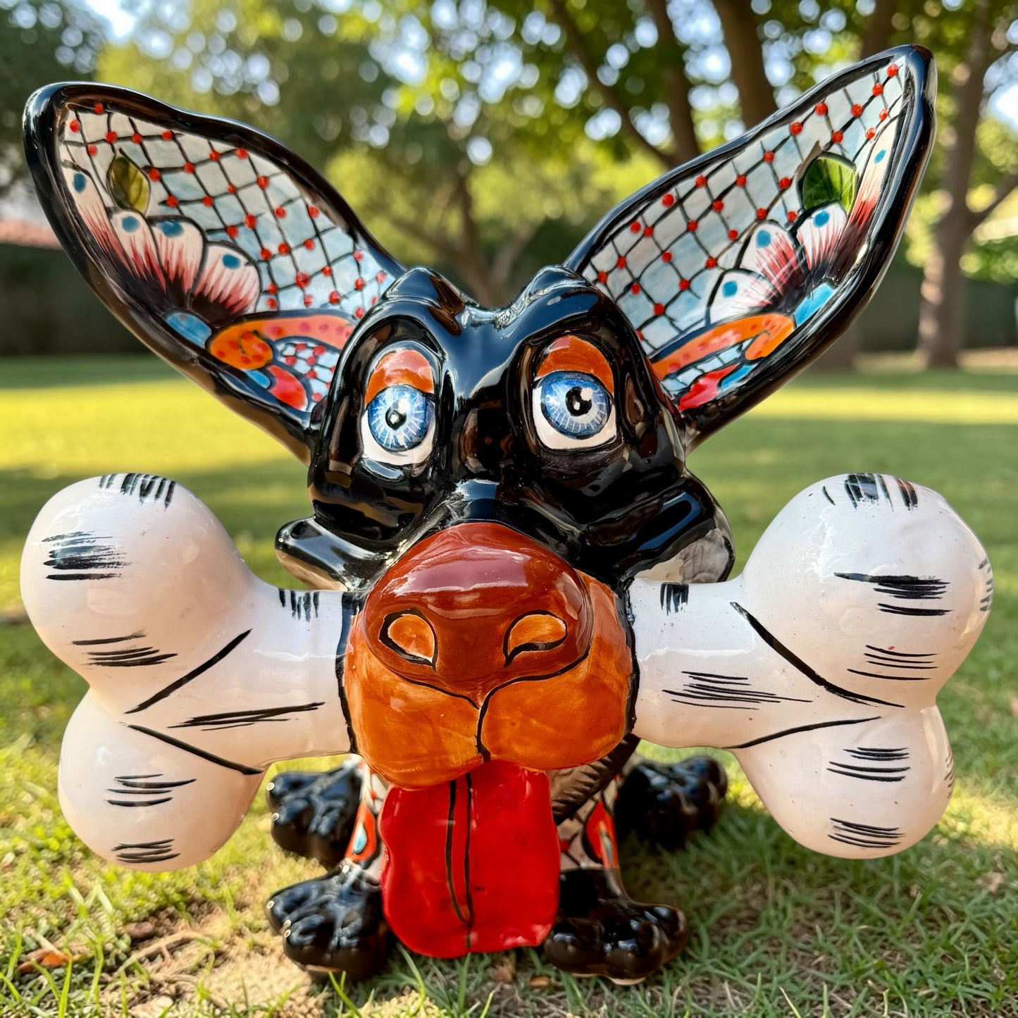 A colorful Talavera dog statue holding a big white bone, photographed outdoors on grass with trees in the background. The dog has a glossy black body, blue eyes, an orange snout, and multicolored patterned ears. 
