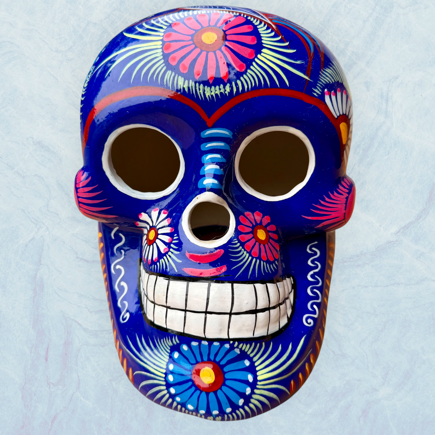 A glossy blue Day of the Dead sugar skull XGrande decorated with vibrant pink, blue, white, and orange floral and geometric patterns, shown against a light blue textured background, front face view.