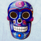 A glossy blue Day of the Dead sugar skull XGrande decorated with vibrant pink, blue, white, and orange floral and geometric patterns, shown against a light blue textured background, front face view.