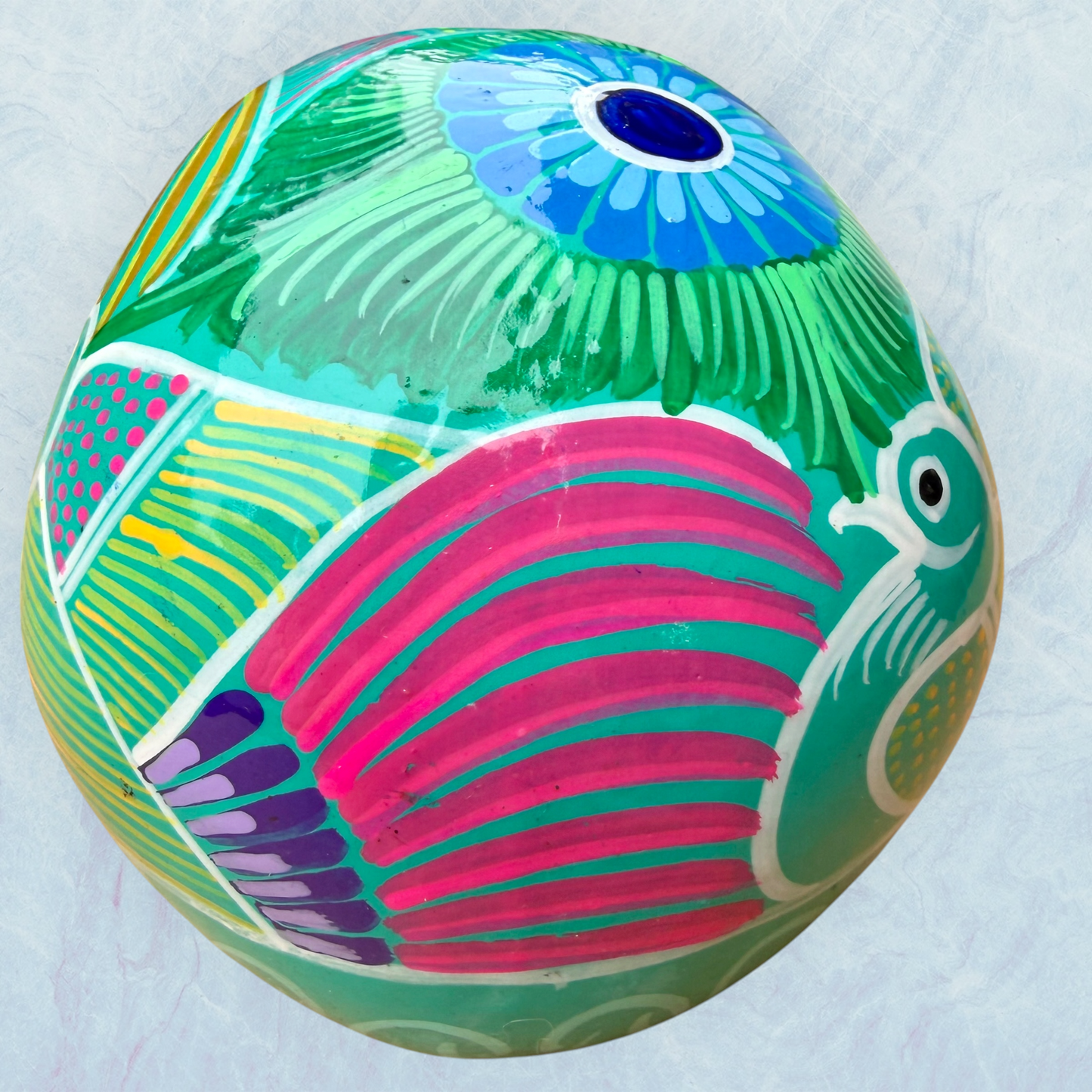 A decorative skull sculpture with intricate colorful sugar skull style patterns painted on a teal colored base, set against a light blue textured background, back view.