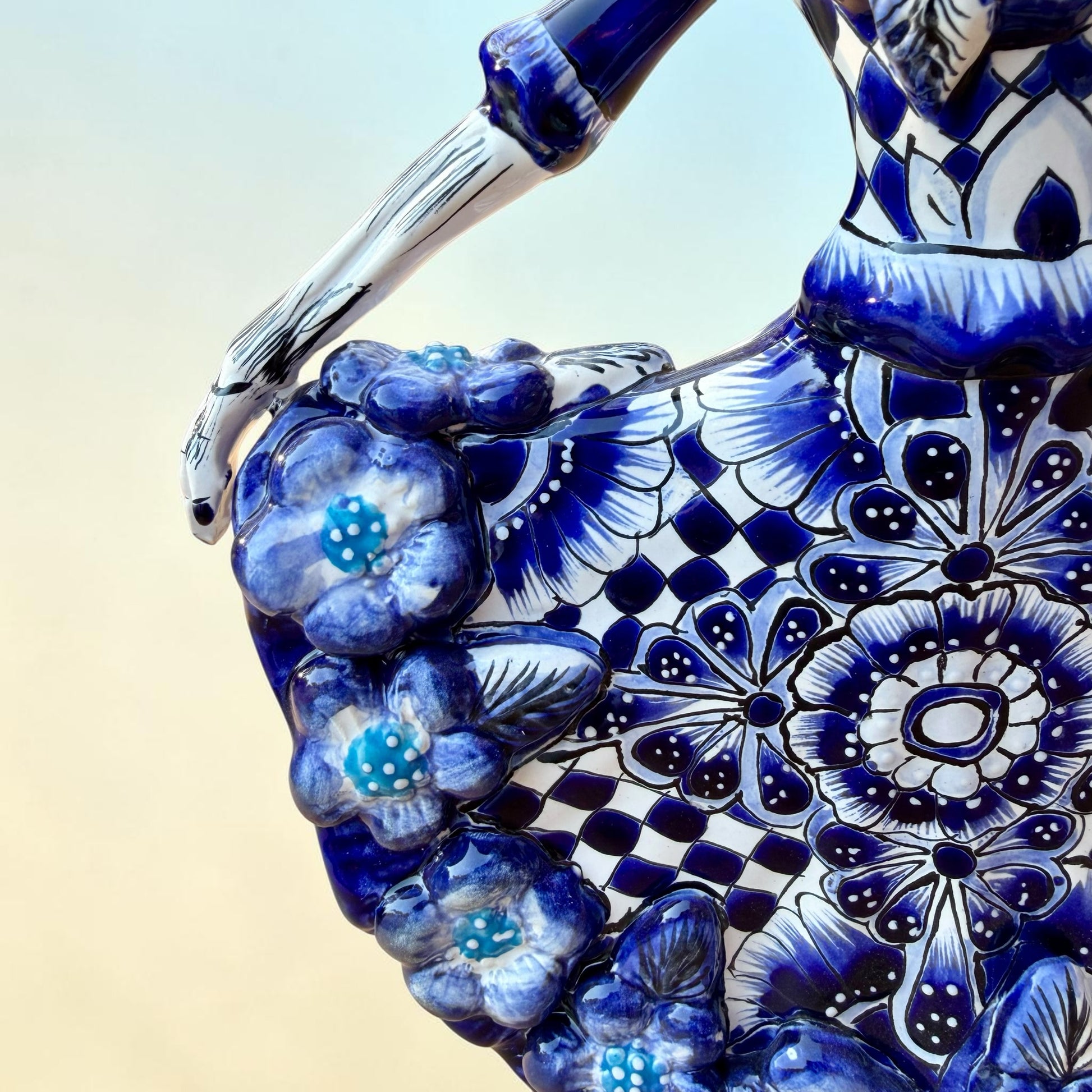 A Day of the Dead style talavera figurine of a skeleton woman in a wide floral skirt, painted in deep blue, black, white, and light blue on a soft beige-to-blue gradient background. Close-up of dress detail.