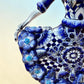 A Day of the Dead style talavera figurine of a skeleton woman in a wide floral skirt, painted in deep blue, black, white, and light blue on a soft beige-to-blue gradient background. Close-up of dress detail.