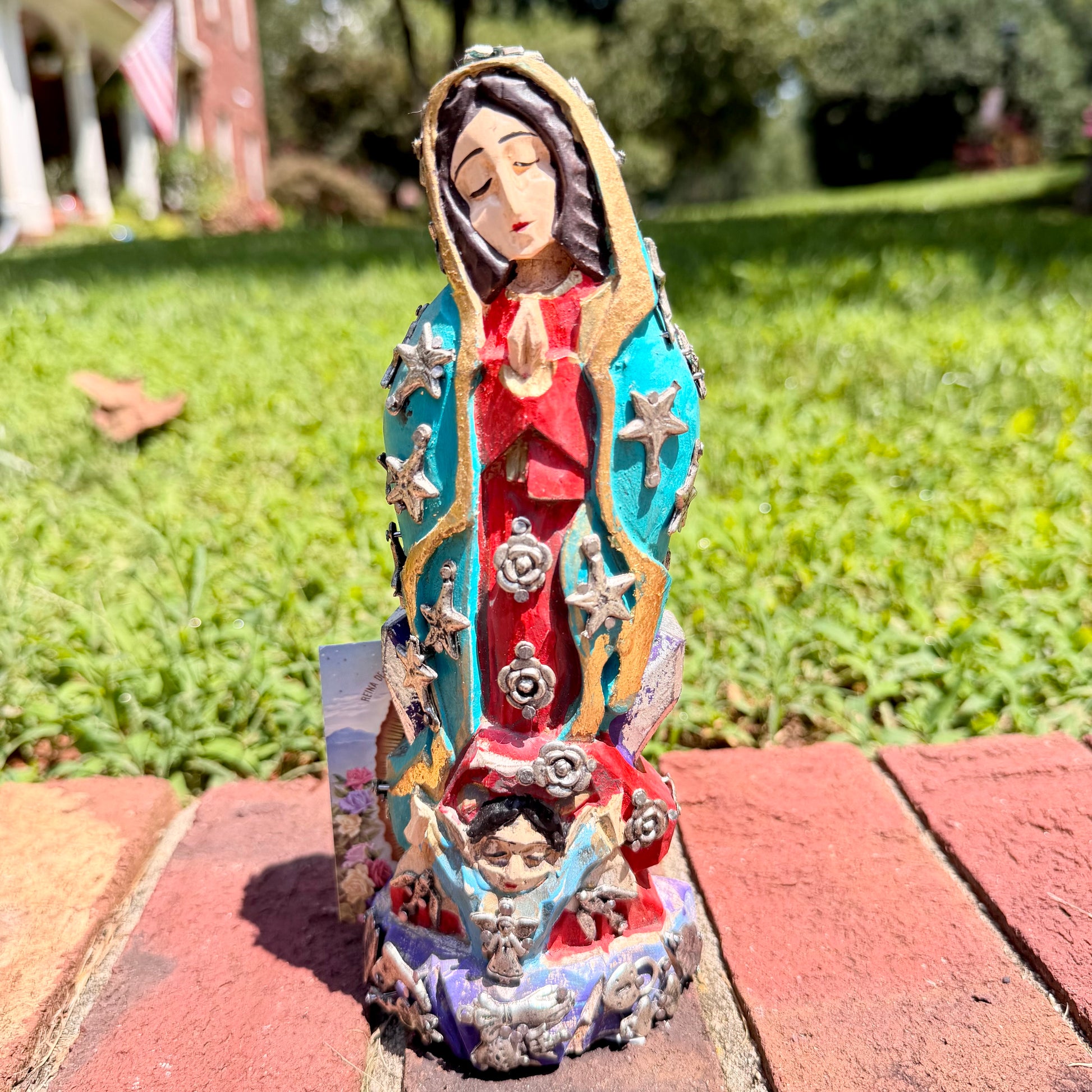 A colorful Milagros Guadalupe  (Virgin Mary) standing on a brick surface outdoors with a grassy yard in the background.