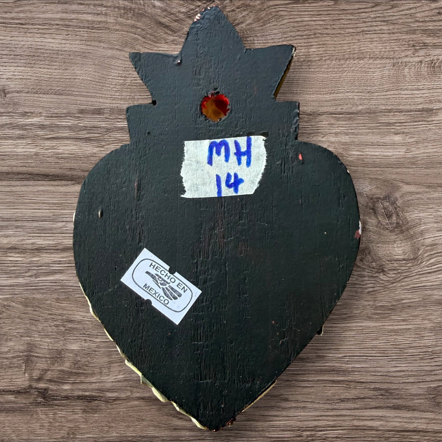 A Milagros heart featuring a gold textured top and a black heart base covered in raised silver-toned charms displayed on a wooden wall, back view. 