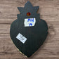 A Milagros heart featuring a gold textured top and a black heart base covered in raised silver-toned charms displayed on a wooden wall, back view. 