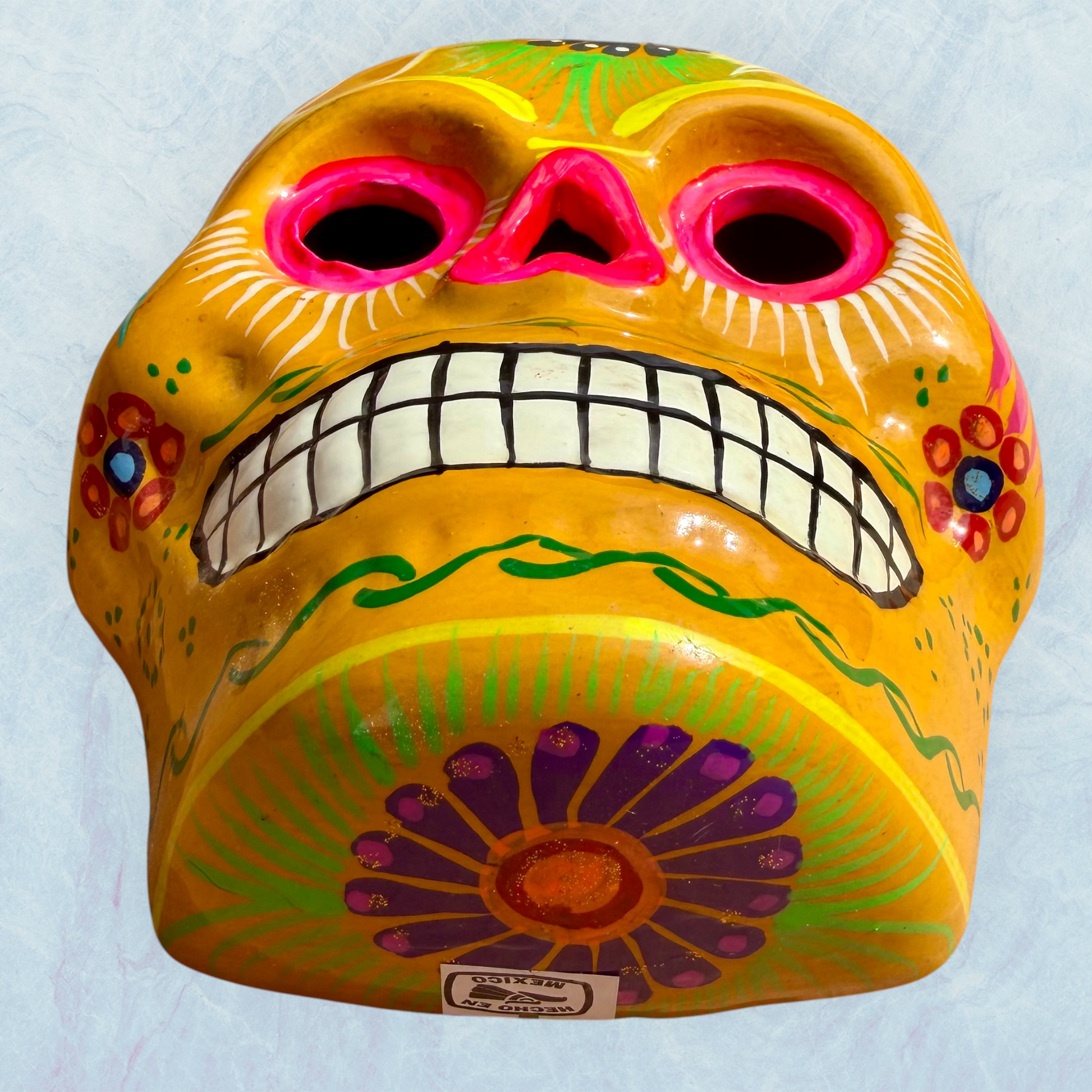 A clay, yellow with floral designs, Mexican-style skeleton skull face mask hanging on a soft blue surface, bottom chin view showing floral.