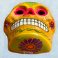 A clay, yellow with floral designs, Mexican-style skeleton skull face mask hanging on a soft blue surface, bottom chin view showing floral.