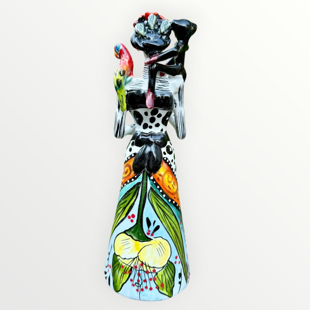 A Day of the Dead Catrina of a Frida skeleton doll holding a black cat, with a red parrot on her shoulder and colorful flowers around the top of her dress. Back view showing the back of her dress with a bow. 