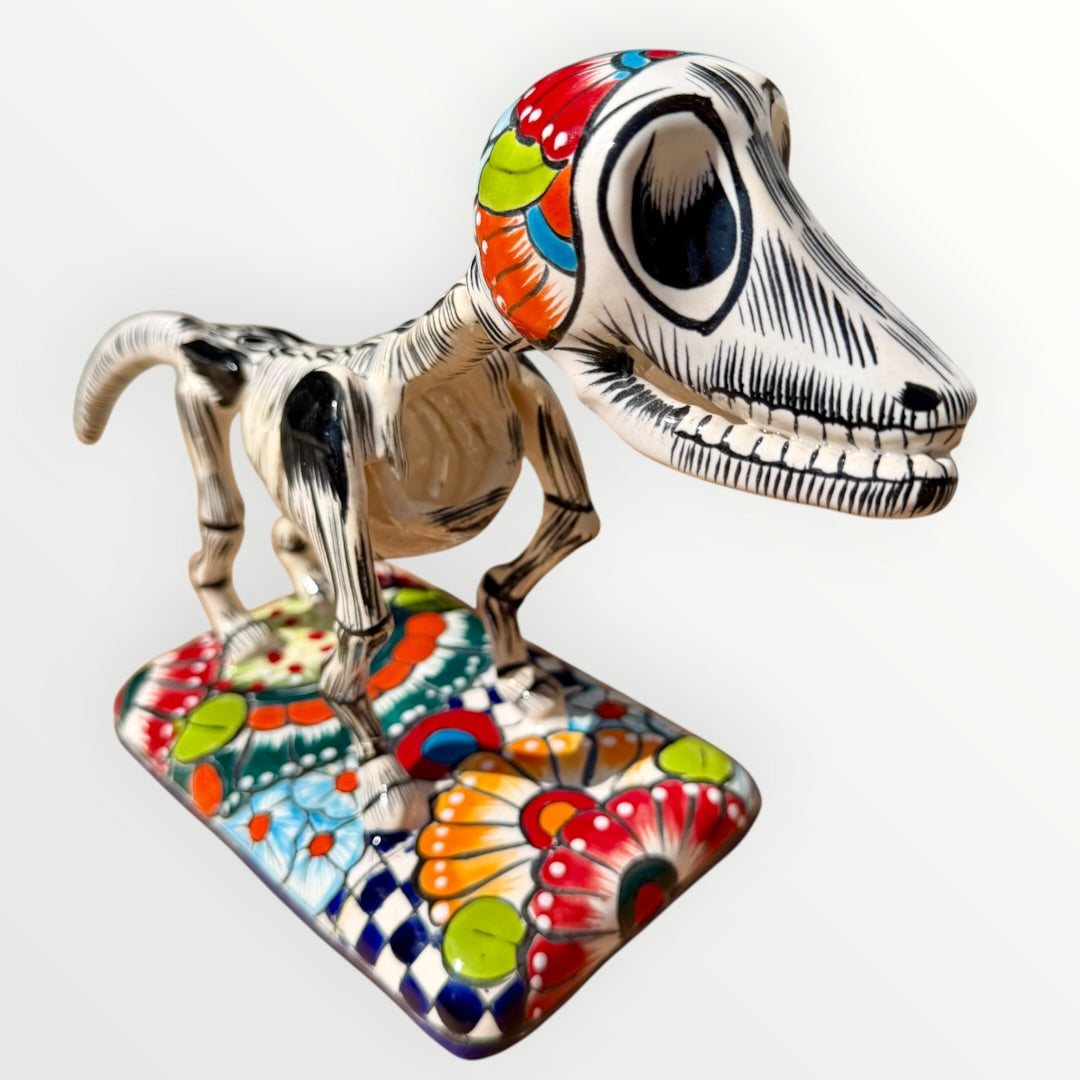 A ceramic Mexican-style skeleton dog figurine standing on a colorful floral base against a plain white background.