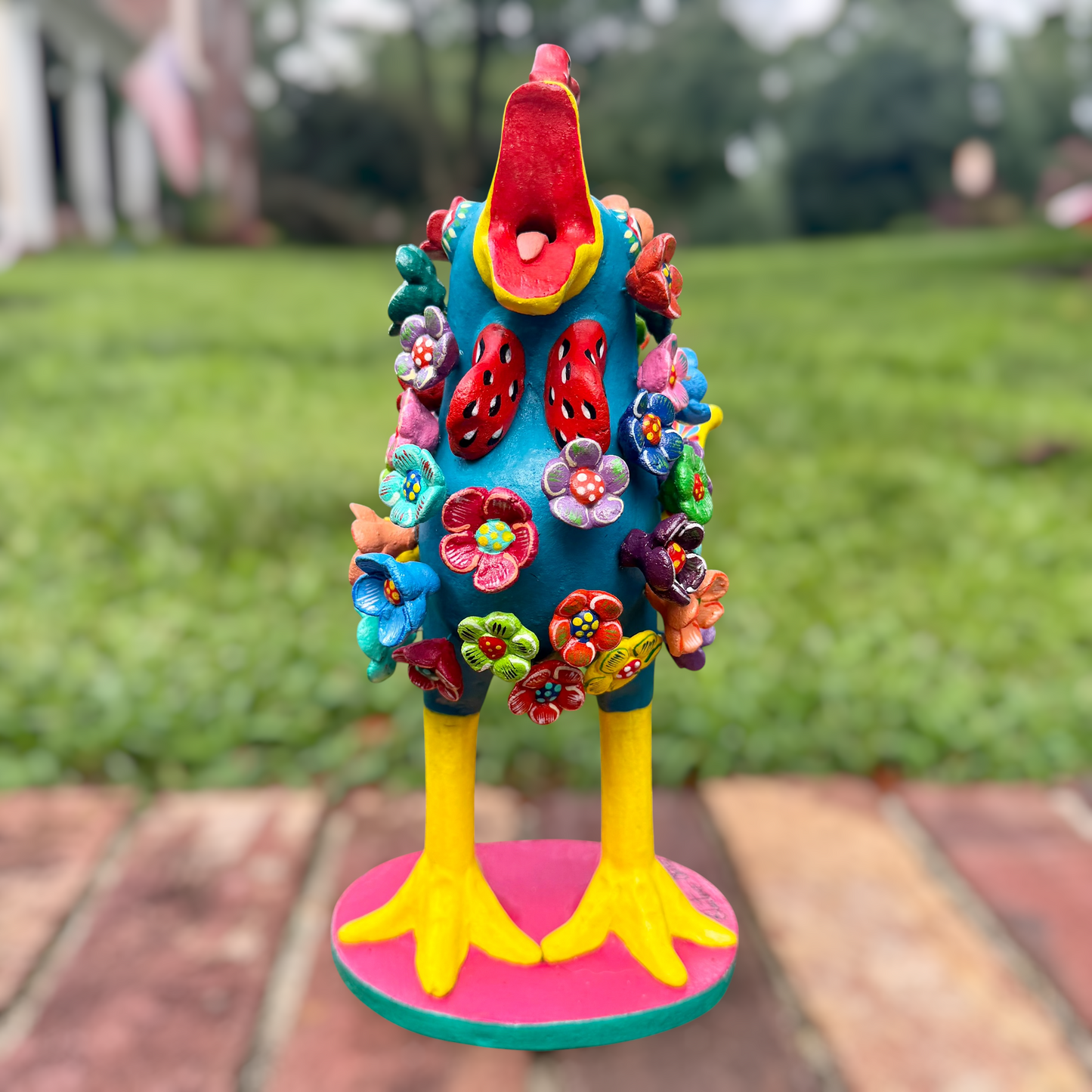 A colorful decorative rooster folk art sculpture with a blue body, yellow legs and beak, and many bright flower ornaments, standing on a pink circular base outdoors on a brick path with a blurred green lawn behind it. Front view with open beak. 