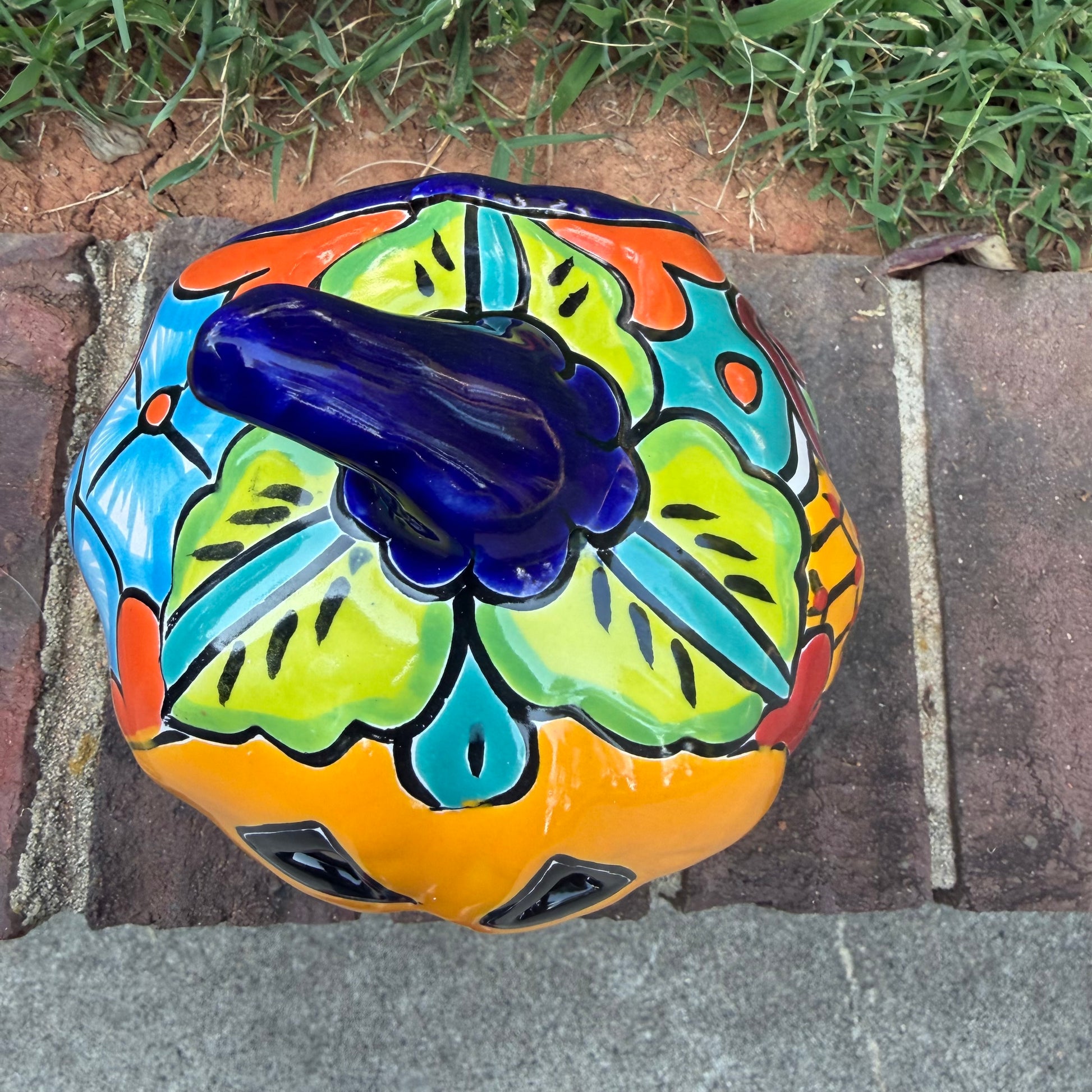 A Talavera jack-o'-lantern pumpkin decoration with a carved face, sitting on a brick surface in an outdoor yard with grass and trees in the background. Top view showing stem on pumpkin.