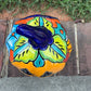 A Talavera jack-o'-lantern pumpkin decoration with a carved face, sitting on a brick surface in an outdoor yard with grass and trees in the background. Top view showing stem on pumpkin.