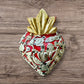 A Mexican Milagros heart in red with a gold flaming top, covered in antiqued charms. 