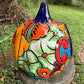 A hand-painted ceramic jack-o'-lantern pumpkin ornament with a bright orange face, red carved eyes and mouth, a blue stem, and colorful side patterns, sitting outdoors on a stone pedestal in a grassy yard. This is the right side view.