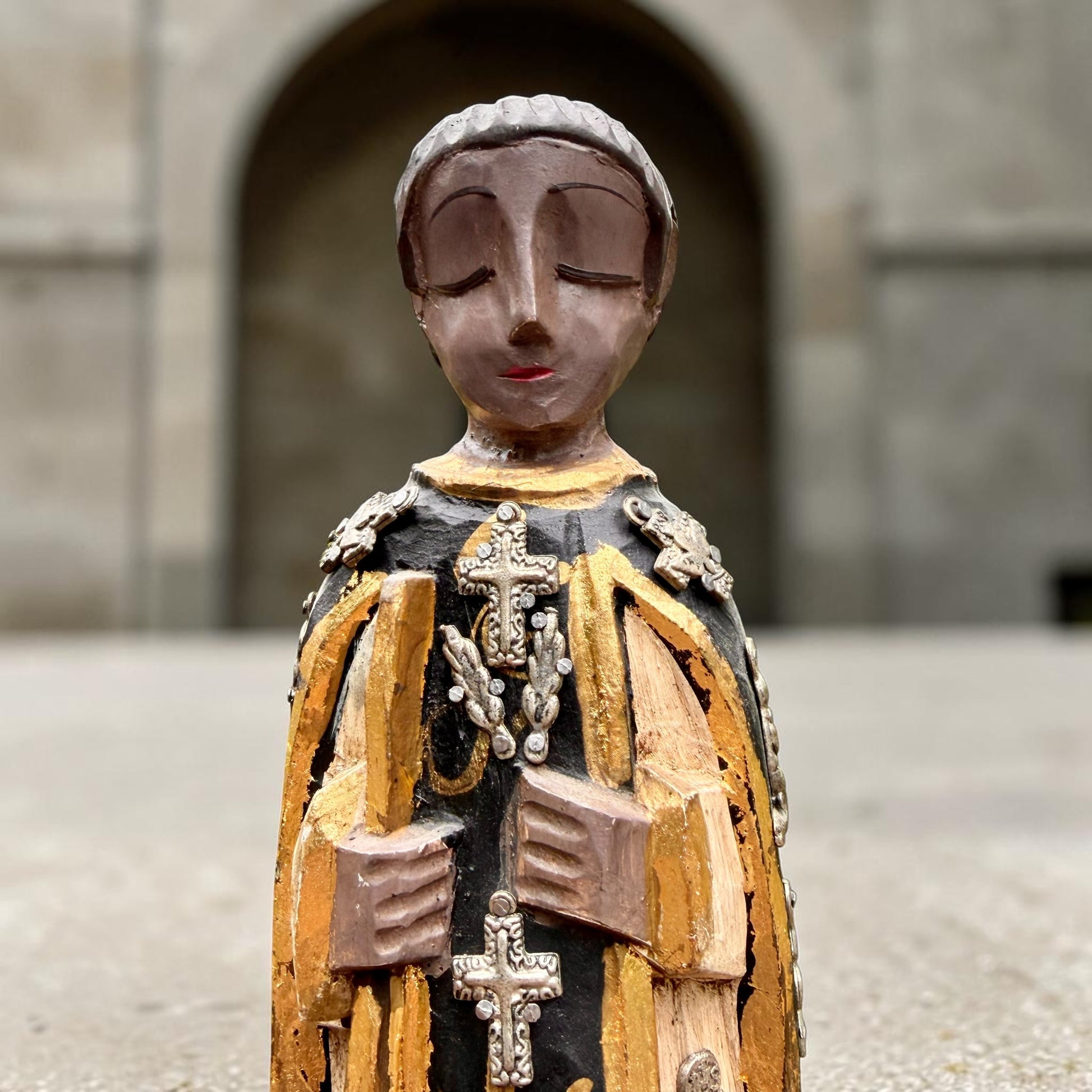 A carved milagros religious figurine of  Saint Martin in ornate black and gold vestments with silver cross details, photographed outdoors in front of a blurred stone archway.