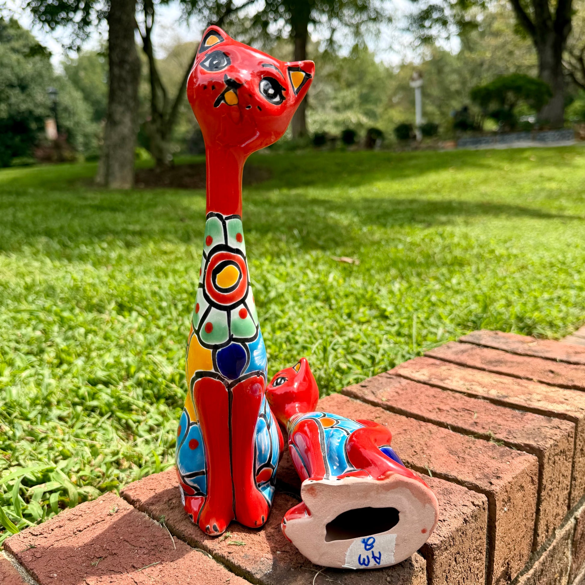 A colorful hand-painted ceramic cat figurine stands on a brick ledge in a grassy outdoor garden, with a second cat figurine lying beside it.