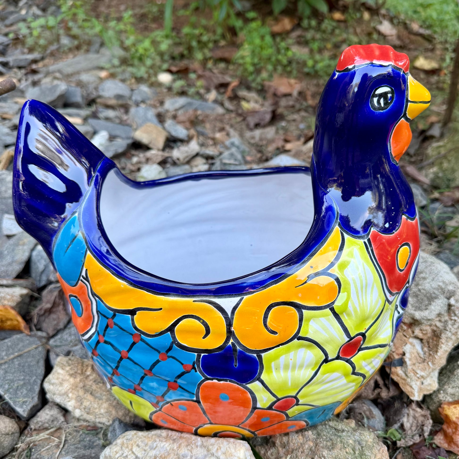 A colorful ceramic chicken-shaped planter with a glossy hand-painted finish, placed outdoors on a bed of small gray rocks and greenery, left side view.