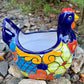 A colorful ceramic chicken-shaped planter with a glossy hand-painted finish, placed outdoors on a bed of small gray rocks and greenery, left side view.