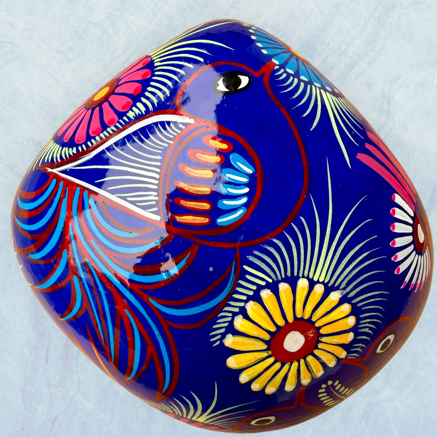 A glossy blue Day of the Dead sugar skull XGrande decorated with vibrant pink, blue, white, and orange floral and geometric patterns, shown against a light blue textured background, top view of skull.