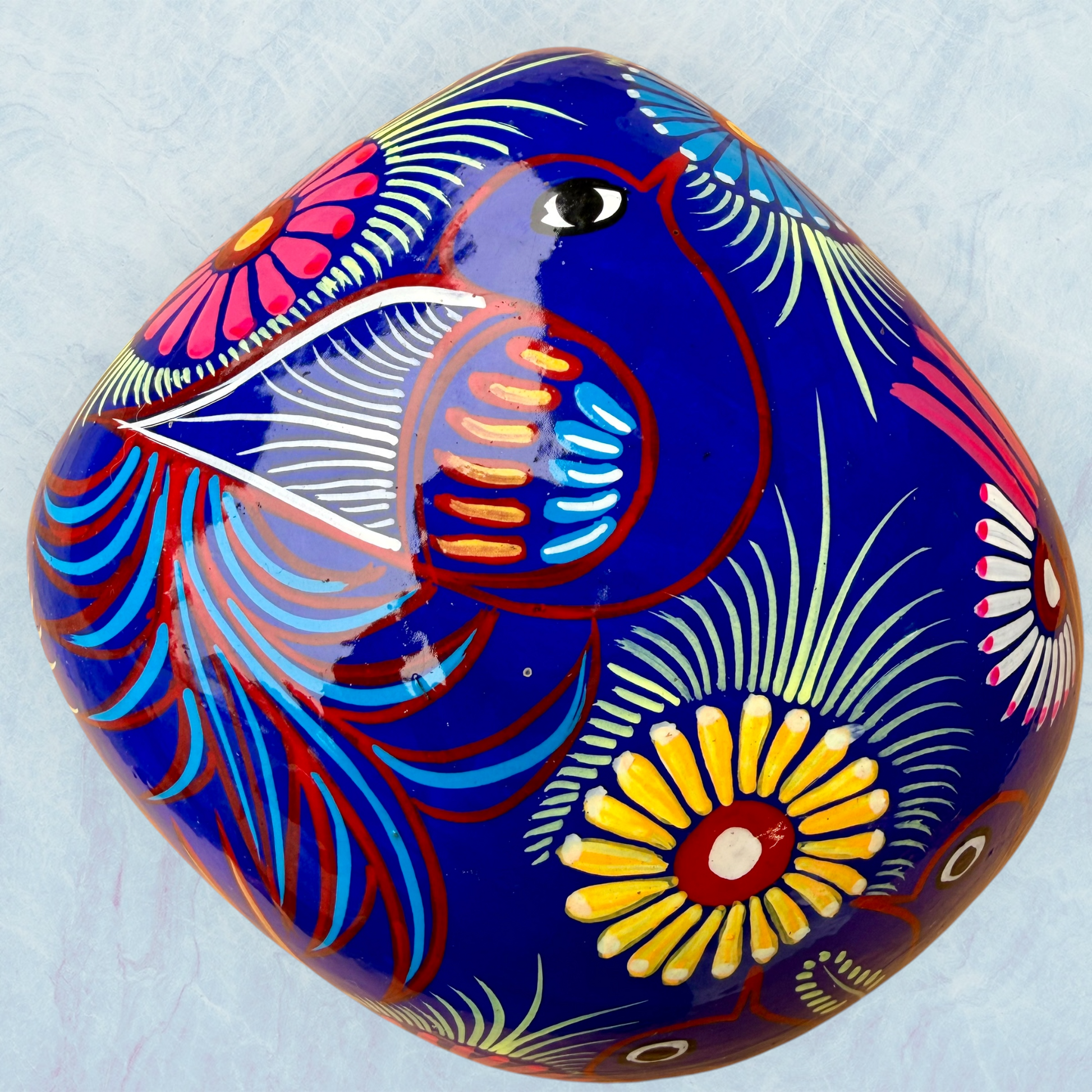 A glossy blue Day of the Dead sugar skull XGrande decorated with vibrant pink, blue, white, and orange floral and geometric patterns, shown against a light blue textured background, top view of skull.