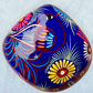 A glossy blue Day of the Dead sugar skull XGrande decorated with vibrant pink, blue, white, and orange floral and geometric patterns, shown against a light blue textured background, top view of skull.