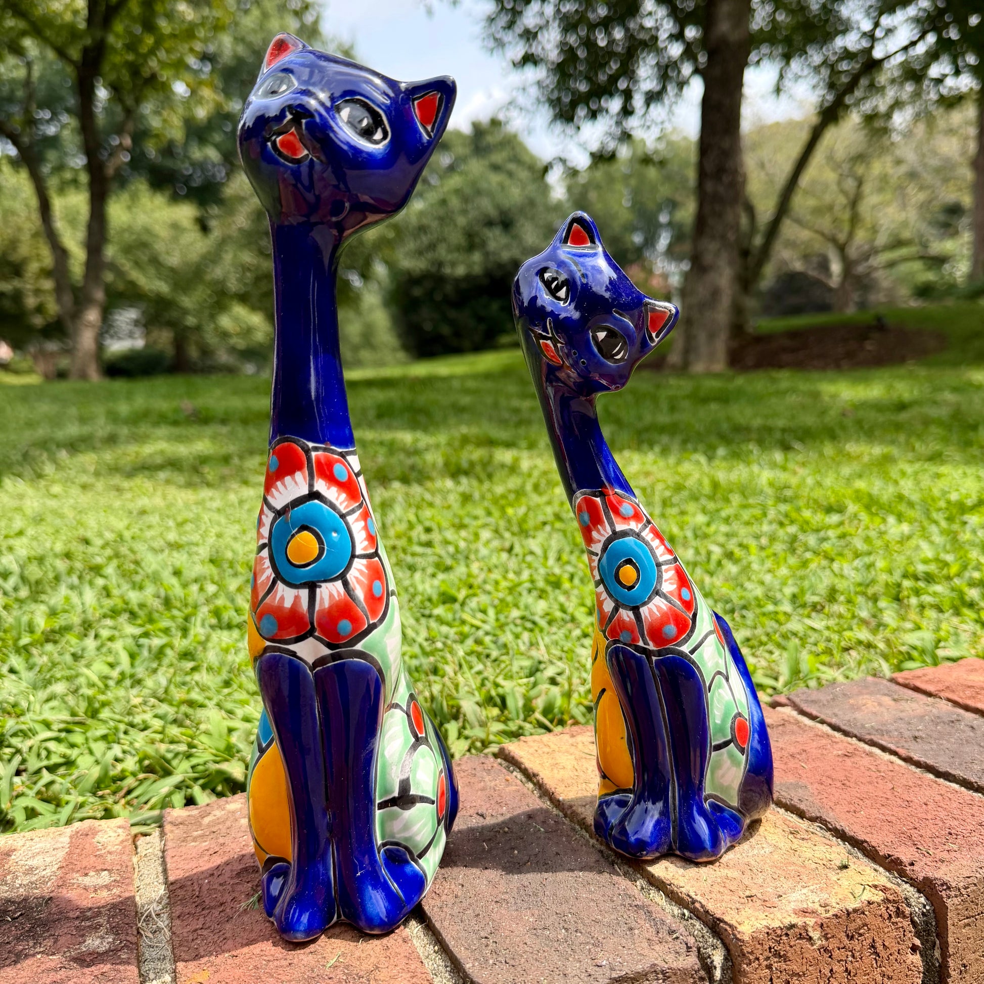 Two colorful glazed ceramic cat figurines sitting on a brick path in a grassy outdoor garden setting.