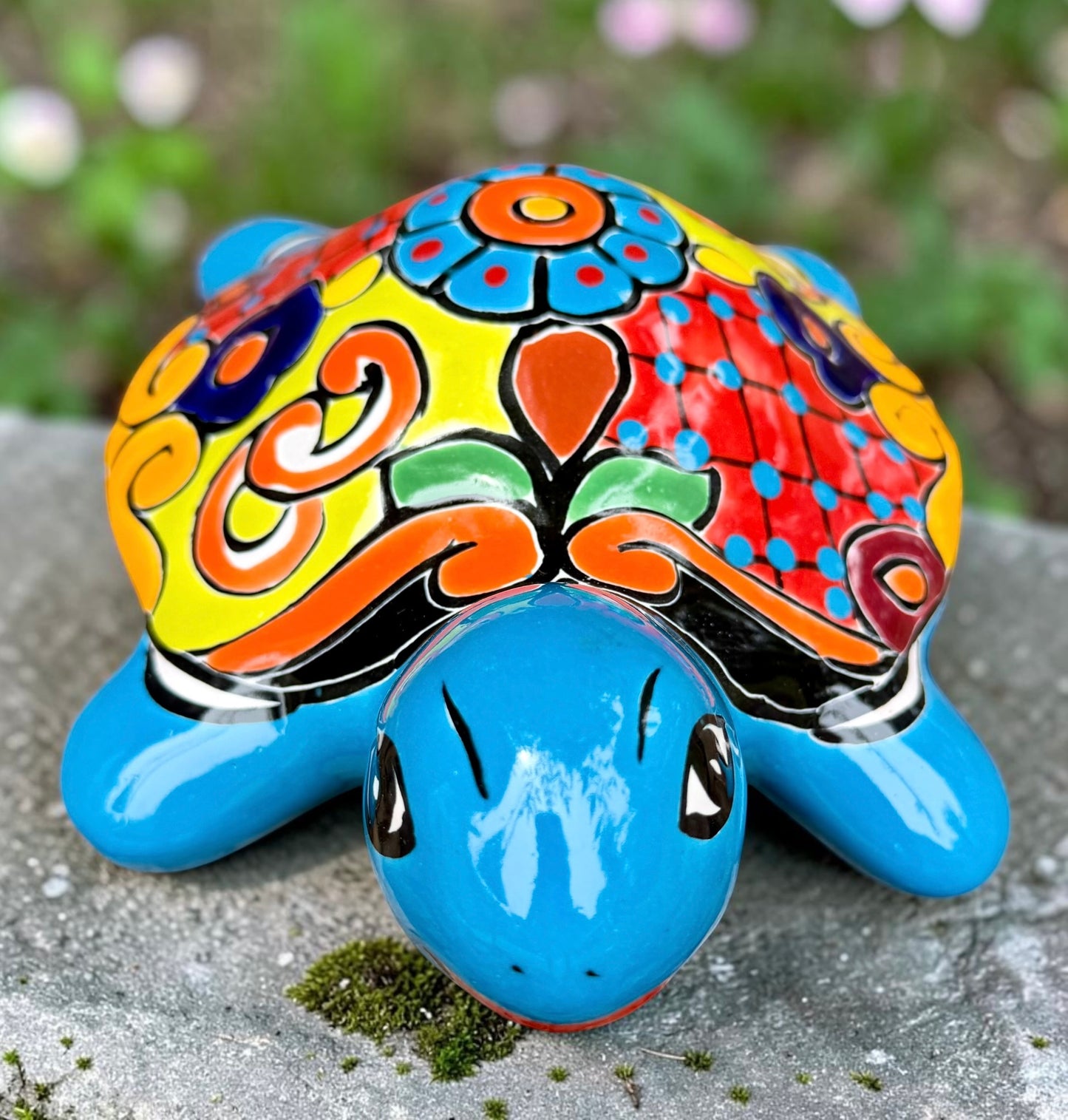 A hand-painted ceramic turtle figurine resting on a mossy stone outdoors, featuring a bright blue body and a highly decorated shell with vibrant orange, yellow, red, and blue patterns, front view.