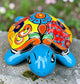 A hand-painted ceramic turtle figurine resting on a mossy stone outdoors, featuring a bright blue body and a highly decorated shell with vibrant orange, yellow, red, and blue patterns, front view.